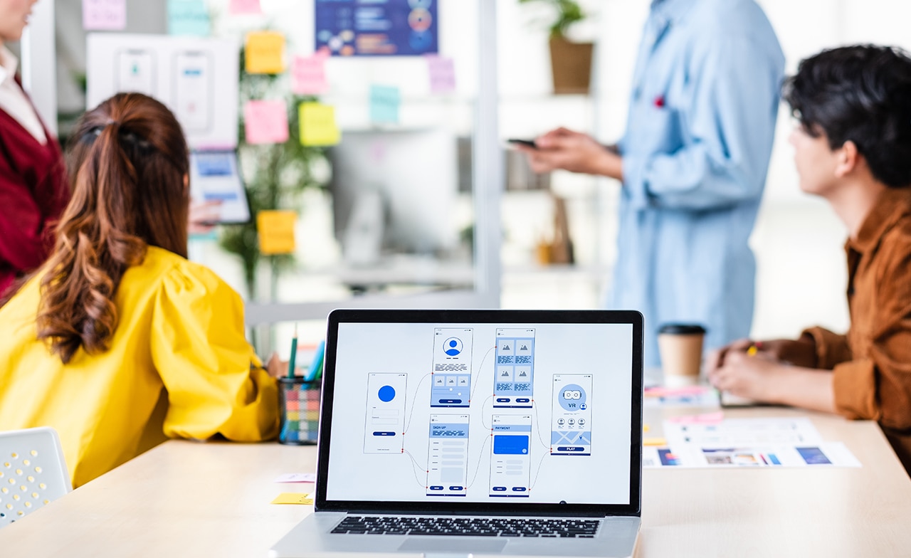 Close up of a laptop with images showing development of a mobile app and a team working in the background on tablets and with post it notes.