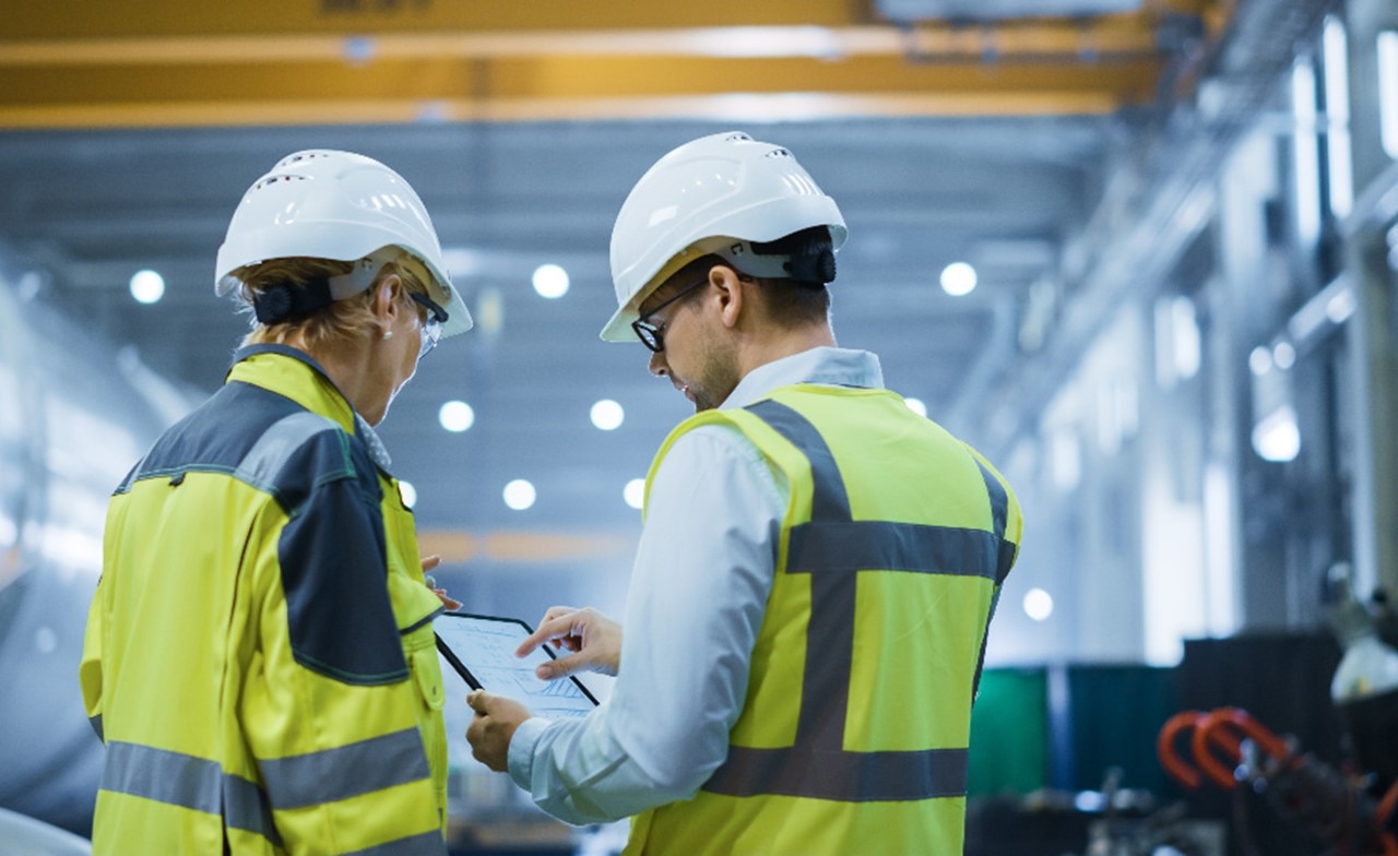 Two individuals in safety vests and helmets inside an industrial facility, one holding a clipboard.