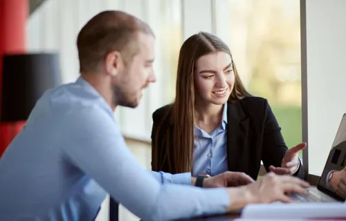 Två kollegor sitter tillsammans vid en bärbar dator och diskuterar, den ena visar något på…
