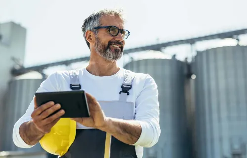 Mitarbeiter in Arbeitskleidung steht vor Industrieanlagen und arbeitet mit einem Tablet.